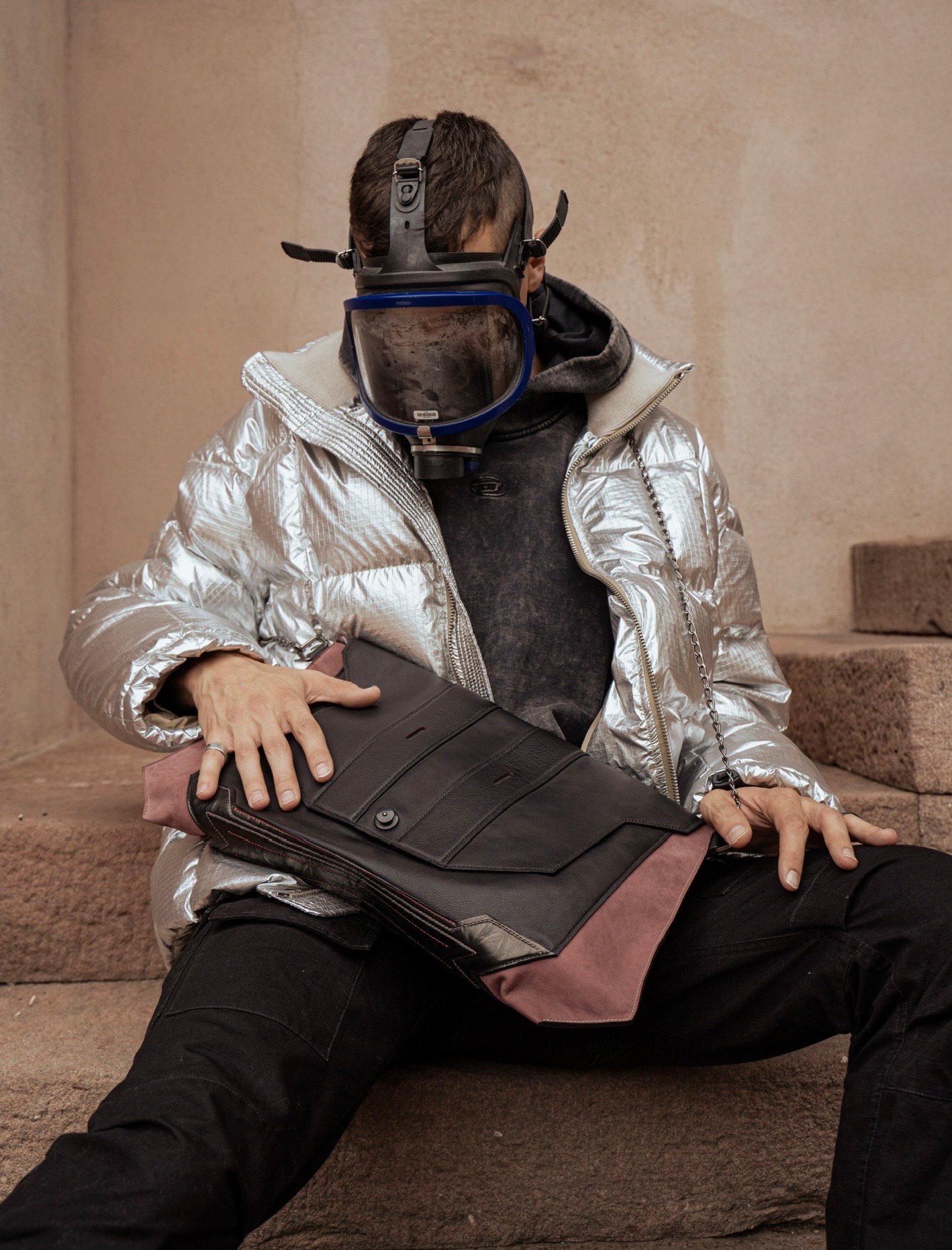 Silhouette assise sur des marches de pierre, veste argentée et masque intégral, tenant contre elle une grande pochette en cuir noir et daim rose