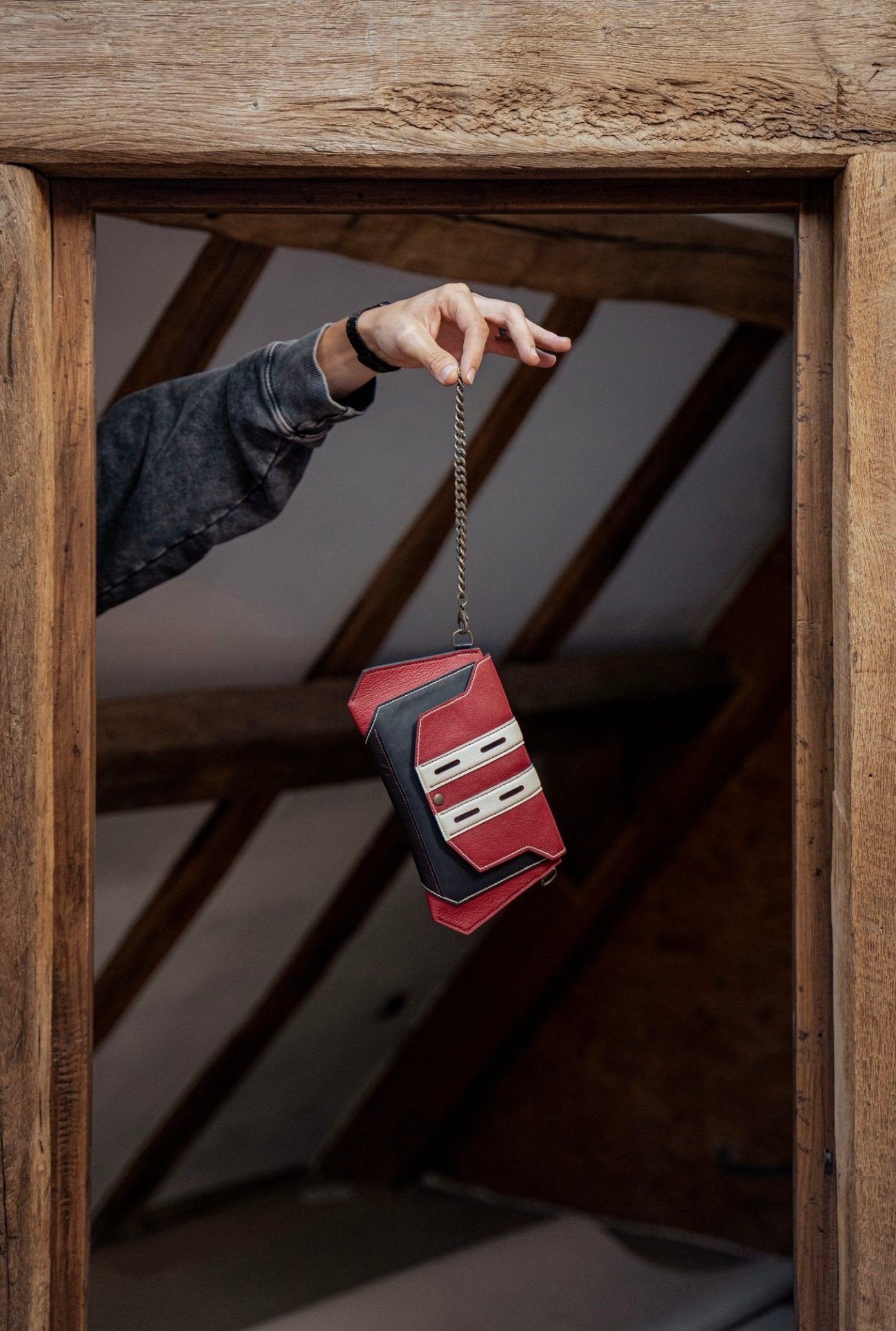 Petite sacoche en cuir tricolore — rouge, noir et écru — suspendue à sa chaîne et tenue à la main, dans l'atelier aux poutres de bois